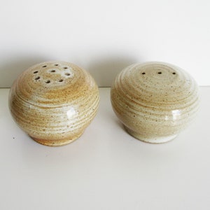 May include: A pair of ceramic salt and pepper shakers. The shakers are spherical with a speckled, tan glaze and a ringed texture. The salt shaker has multiple holes, while the pepper shaker has three.