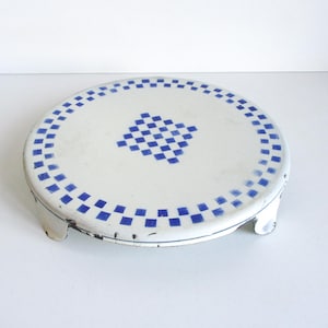 Puede incluir: Un soporte redondo de metal blanco para pasteles con un patrón de cuadros azules en la superficie superior y alrededor del borde. El soporte tiene tres patas cortas y curvas. Diseño vintage y decorativo.
