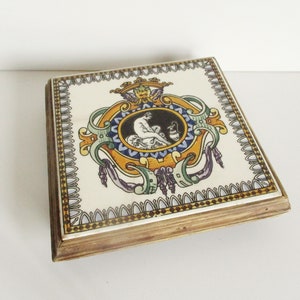 May include: Square decorative box with a ceramic top featuring a detailed emblem in a gold, blue, orange, and purple color scheme. The box has a light brown wooden frame and a white background.