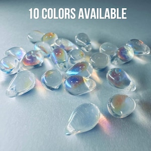 Puede incluir: Una colección de pequeñas cuentas de vidrio transparentes en forma de lágrima con un acabado iridiscente que refleja colores del arcoíris. El texto 10 COLORS AVAILABLE está impreso en blanco en la parte superior, indicando la variedad.