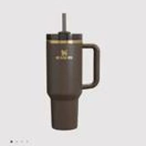 May include: A dark brown insulated tumbler with a matching handle, a black lid, and a central straw. The front features a gold-colored logo and the text STANLEY. The cylindrical body tapers at the base, designed for standard cup holders, with a matte finish.