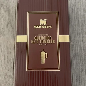 May include: A rectangular dark maroon product box for a Stanley The Flowstate Quencher H2.0 Tumbler. The box features gold text and a gold logo, with the capacity clearly printed as 40 oz and 1.18 L, along with a graphic of the tumbler.