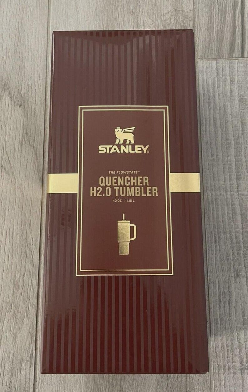 May include: A rectangular dark maroon product box for a Stanley The Flowstate Quencher H2.0 Tumbler. The box features gold text and a gold logo, with the capacity clearly printed as 40 oz and 1.18 L, along with a graphic of the tumbler.