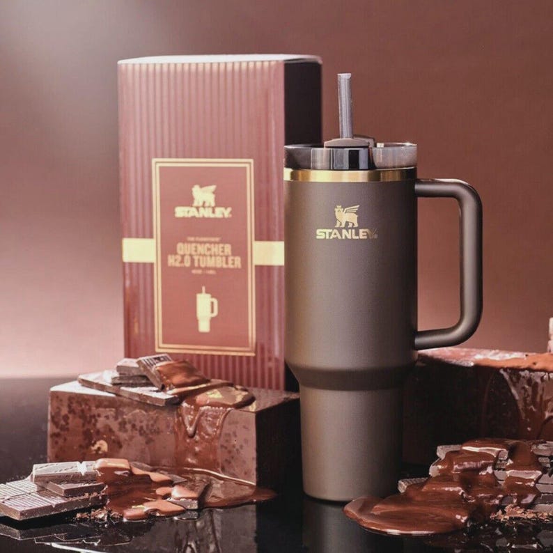 May include: A dark brown Stanley Quencher H2.0 tumbler with a matching handle, a gold-tone rim, and a straw. The tumbler features the Stanley logo in gold. It is displayed next to its original box and several blocks of melting dark chocolate.
