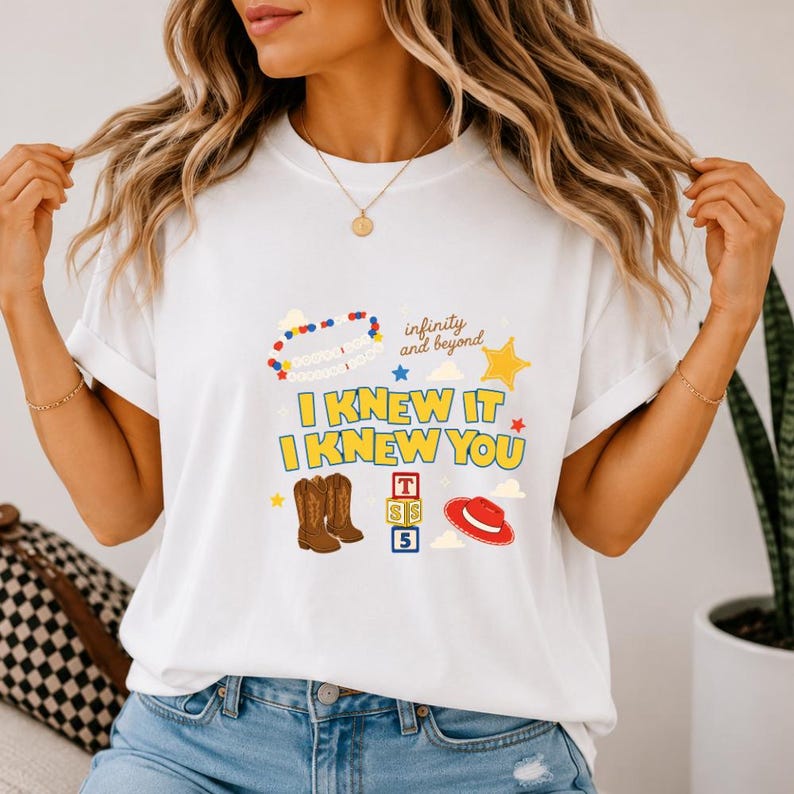 May include: A white short-sleeved t-shirt featuring a graphic print with the text "I KNEW IT I KNEW YOU" and "infinity and beyond". The design includes illustrations of a beaded necklace, a yellow star, brown cowboy boots, alphabet blocks, and a red hat.