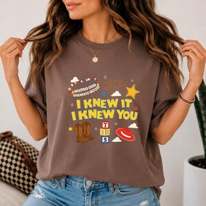 May include: A brown short-sleeve t-shirt featuring a graphic print with the text "I KNEW IT I KNEW YOU" in yellow, "YOU'VE GOT A FRIEND IN ME" in a circular pattern, and "Infinity and beyond". Includes illustrations of cowboy boots, blocks, and a hat.
