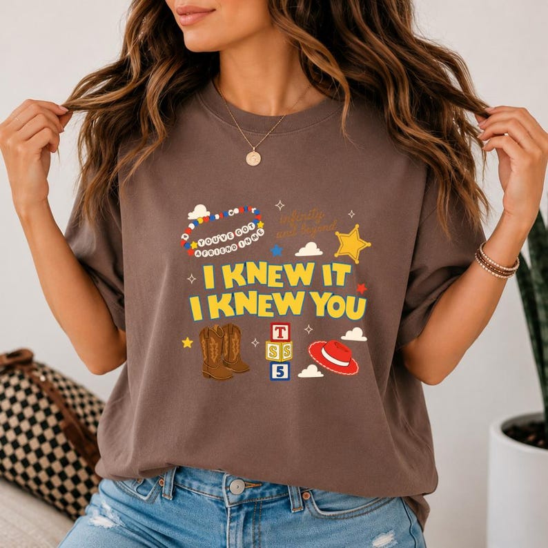 May include: A brown short-sleeve t-shirt featuring a graphic print with the text "I KNEW IT I KNEW YOU" in yellow, "YOU'VE GOT A FRIEND IN ME" in a circular pattern, and "Infinity and beyond". Includes illustrations of cowboy boots, blocks, and a hat.