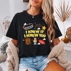 May include: A black short-sleeve t-shirt featuring a graphic print with the text "I KNEW IT I KNEW YOU", "YOU'VE GOT A FRIEND IN ME", and "infinity and beyond". The design includes illustrations of brown cowboy boots, a red hat, and blocks.