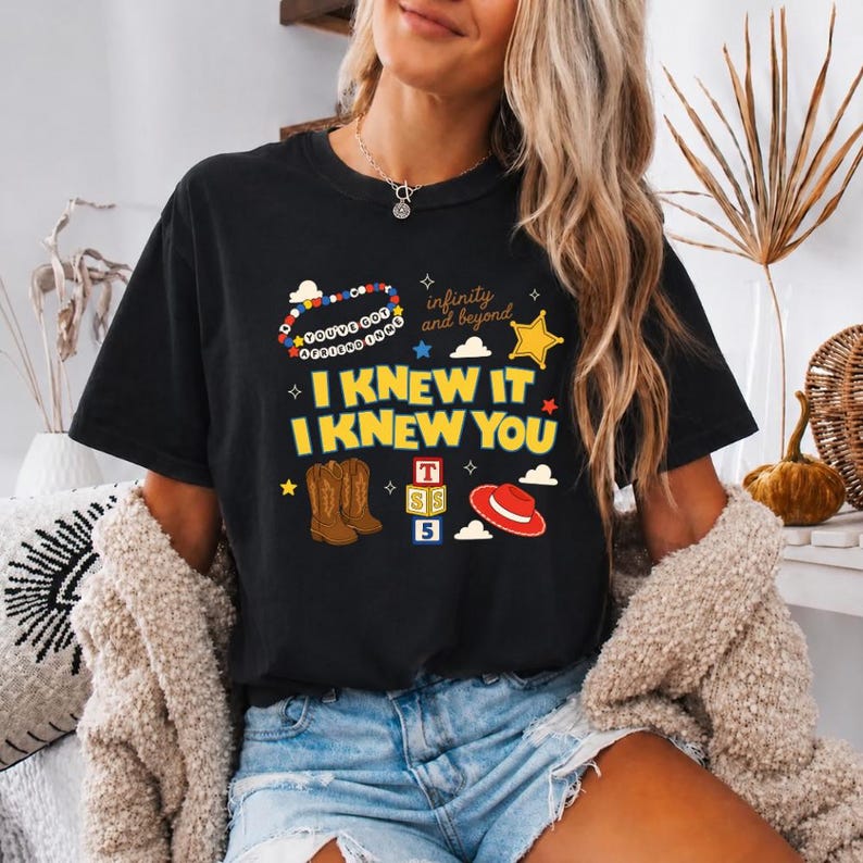 May include: A black short-sleeve t-shirt featuring a graphic print with the text "I KNEW IT I KNEW YOU", "YOU'VE GOT A FRIEND IN ME", and "infinity and beyond". The design includes illustrations of brown cowboy boots, a red hat, and blocks.