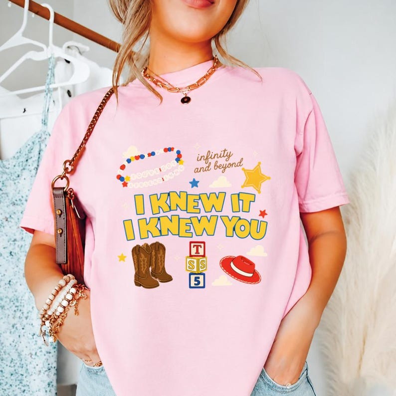May include: A light pink t-shirt featuring a graphic print with the text "I KNEW IT I KNEW YOU" and "infinity and beyond". The design includes illustrations of a beaded bracelet, a yellow star, brown cowboy boots, alphabet blocks, and a red hat.