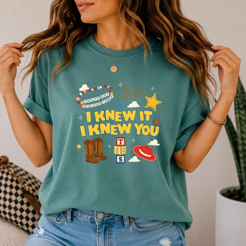 May include: A teal t-shirt featuring a graphic print with the text I KNEW IT I KNEW YOU, a beaded bracelet graphic with the text YOU'VE GOT A FRIEND IN ME, brown cowboy boots, a red cowboy hat, a yellow sheriff star, and stacked alphabet blocks.