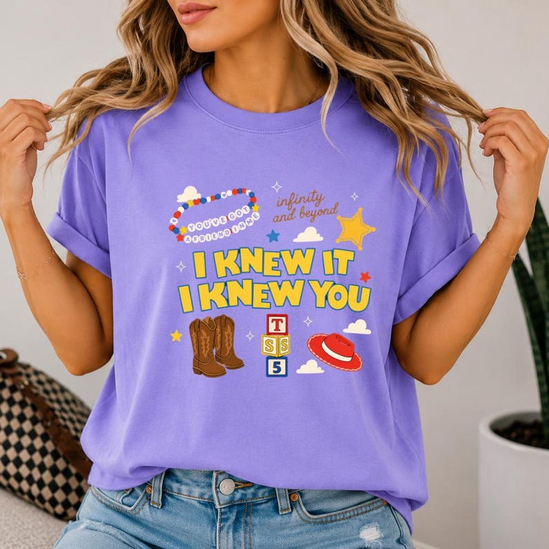 May include: A light purple short-sleeve t-shirt featuring a graphic print with the text "I KNEW IT I KNEW YOU", "infinity and beyond", and "YOU'VE GOT A FRIEND IN ME". The design includes a beaded necklace, cowboy boots, alphabet blocks, and a hat.