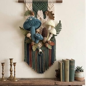 May include: A macrame wall hanging featuring a wooden dowel, cream and dark green knotted cord, and crocheted mushrooms in blue, white, and brown. The piece is decorated with crocheted fern leaves, oak leaves, and small acorns attached to the center.