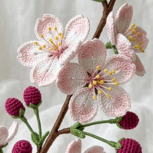 May include: A handcrafted crochet cherry blossom branch featuring light pink and white five-petaled flowers with yellow centers, dark magenta buds, and green stems attached to a brown branch. The intricate yarn work creates a realistic floral display.