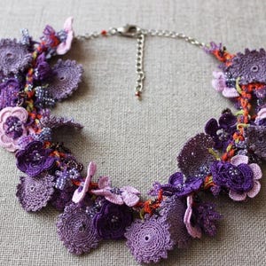 Peut inclure: Un collier composé d'un arrangement dense de motifs floraux au crochet dans diverses nuances de violet, lavande et pourpre. Le design intègre de petites perles de verre et des accents de fil orange, fini avec une chaîne en métal argenté.