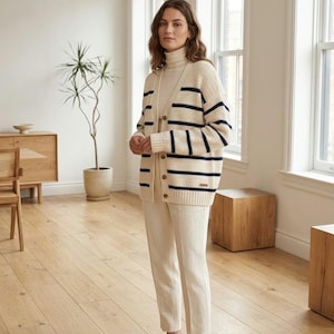 Peut inclure: Un cardigan en tricot couleur crème avec des rayures horizontales bleu marine, un col en V et une fermeture boutonnée à l'avant avec des boutons marron. Le vêtement est porté sur un pull à col roulé et un pantalon assortis couleur crème.