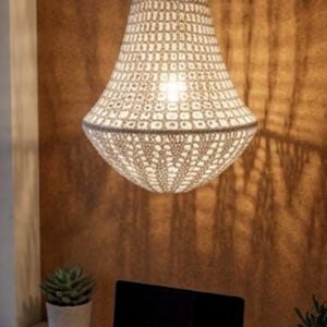 May include: A white crocheted pendant light fixture with a bell-shaped shade featuring an intricate open-weave pattern. The lamp hangs from a central cord and emits a warm glow, casting a textured shadow pattern onto the wall behind it.