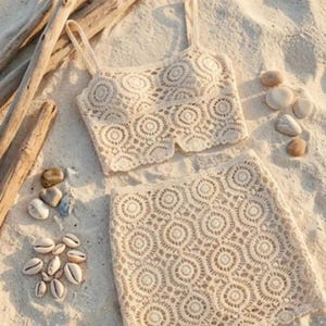 May include: A two-piece set consisting of a cream-colored crochet crop top with thin straps and a matching crochet mini skirt. Both pieces feature a repeating circular pattern with scalloped edges, crafted from a light-colored yarn material.
