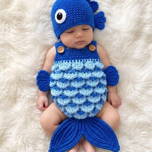 May include: A blue crochet fish costume consisting of a hat with a white and black eye and a dorsal fin, and a romper featuring layered light and dark blue scales, side fins, and a large tail fin at the bottom, secured with two brown wooden buttons.