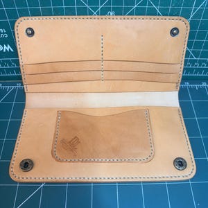 May include: A tan leather bifold wallet featuring multiple card slots, a central pocket, and four metal snap fasteners at the corners. The interior includes a stamped logo reading DL Dixon Leatherworks. The wallet is constructed with visible light-colored stitching.