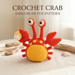 Pode incluir: Um caranguejo amigurumi de crochê com corpo laranja brilhante, barriga amarela e dois talos longos com olhos brancos e pretos. O caranguejo segura uma pequena concha amarela e branca em uma pinça. O texto diz CROCHET CRAB AMIGURUMI PDF PATTERN.