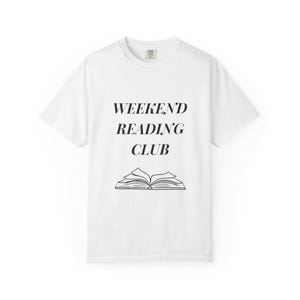以下が含まれることがあります： 胸の中央に黒い文字で「WEEKEND READING CLUB」とプリントされた白い半袖Tシャツ。テキストの下には、ページが見える開いた本のシンプルな黒い線画が描かれています。標準的なクルーネックのTシャツです。
