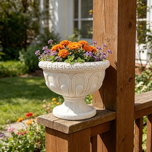 May include: A white stone-look pedestal planter with a rounded bowl featuring embossed vertical arches and a decorative rim, filled with vibrant orange marigolds and small purple flowers, resting on a wooden post in an outdoor setting.