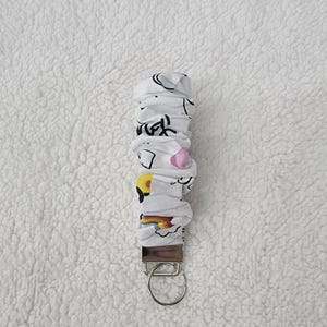 May include: A white fabric keychain wristlet featuring a repeating pattern of colorful cartoon icons including rainbows, yellow shapes, and pink elements. The fabric is gathered onto a silver-toned metal hardware piece with a circular split ring attached.