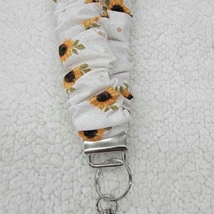 May include: A fabric lanyard featuring a white background with a repeating pattern of yellow sunflowers and green leaves. The fabric is gathered in a scrunchie style and attached to a silver-toned metal clasp and a circular split key ring.