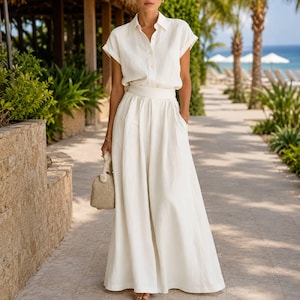 May include: A two-piece set consisting of a short-sleeved, collared button-down shirt and a matching floor-length, high-waisted maxi skirt in an off-white color. The skirt features a wide waistband and a full, flowing silhouette with side pockets.