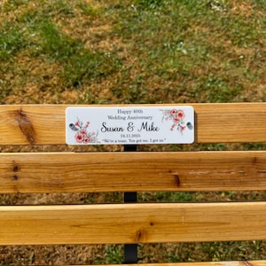 May include: A rectangular white plaque attached to a wooden bench, featuring floral illustrations and the text: Happy 40th Wedding Anniversary, Susan & Mike, 24.11.2025, We're a team. You got me. I got us. The plaque is secured with two silver screws.