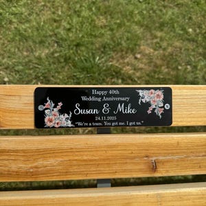 Könnte beinhalten: Ein rechteckiges schwarzes Schild mit floralen Akzenten in den Ecken, das den Text enthält: Happy 40th Wedding Anniversary, Susan & Mike, 24.11.2025, sowie das Zitat: We're a team. You got me. I got us. Das Schild ist mit zwei silbernen Schrauben befestigt.