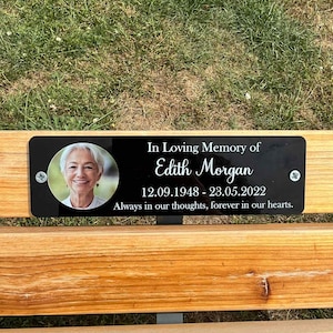 Peut inclure: Une plaque commémorative noire rectangulaire fixée sur un banc en bois, avec un portrait circulaire d'une femme, le texte "In Loving Memory of Edith Morgan", les dates "12.09.1948 - 23.05.2022" et "Always in our thoughts, forever in our hearts."