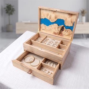 May include: A wooden jewellery box with a hinged lid featuring a blue resin inlay and the text GARVEL. The interior includes a top tray with ring slots and a compartment for necklaces, plus a pull-out drawer with sections for bracelets and earrings.