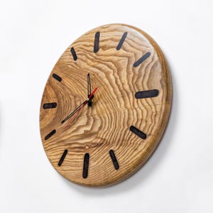 Könnte beinhalten: Eine runde Wanduhr aus hellbraunem Holz mit einer ausgeprägten natürlichen Maserung. Das Zifferblatt verfügt über zwölf dunkle, eingelassene rechteckige Stundenmarkierungen und drei dünne schwarze Zeiger, wobei der Sekundenzeiger rot ist.