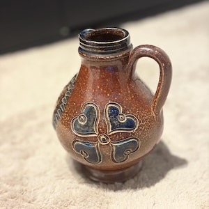 May include: A brown ceramic pitcher with a speckled glaze finish, featuring a single handle and a prominent embossed four-leaf clover design in dark blue. The vessel has a rounded body, a narrow neck, and a flared rim with a small pouring spout.