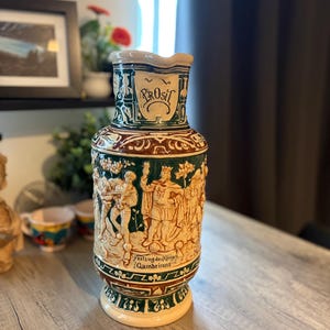 Peut inclure: Une grande carafe à bière en céramique avec un vernis crème et vert foncé, ornée de reliefs détaillés représentant des personnages et des arbres. L'inscription "PROSIT" figure près du col, et la base porte le texte "Festzug des Königs Gambrinus".
