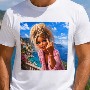Può includere: Una t-shirt bianca a maniche corte con una stampa grafica di una bambola con capelli biondi raccolti in uno chignon spettinato, che indossa un accappatoio rosa e fa il dito medio. Lo sfondo mostra una città costiera sul mare blu.