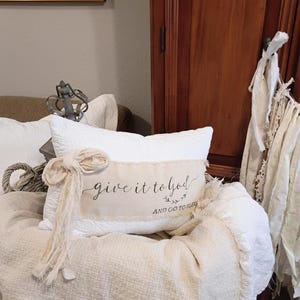 May include: A rectangular white quilted throw pillow featuring a beige fabric strip across the center with the black printed text "give it to god AND GO TO SLEEP" and a decorative frayed fabric bow attached to the left side of the pillow.