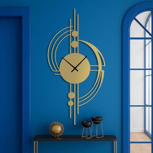 May include: A gold-coloured wall clock featuring a central circular face with black hands, surrounded by an asymmetrical geometric frame of vertical lines, curved arcs, and small circular accents, mounted on a solid blue wall surface.