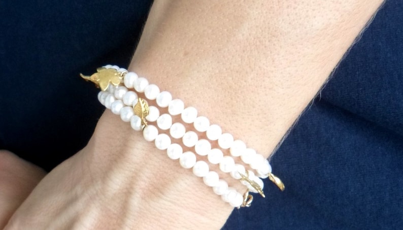 May include: A triple strand pearl bracelet with gold leaf accents. The bracelet is made of white pearls and has a delicate, feminine look.