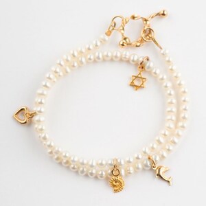 May include: A double strand pearl bracelet with gold charms. The charms include a heart, a star of David, a seashell, and a dolphin.