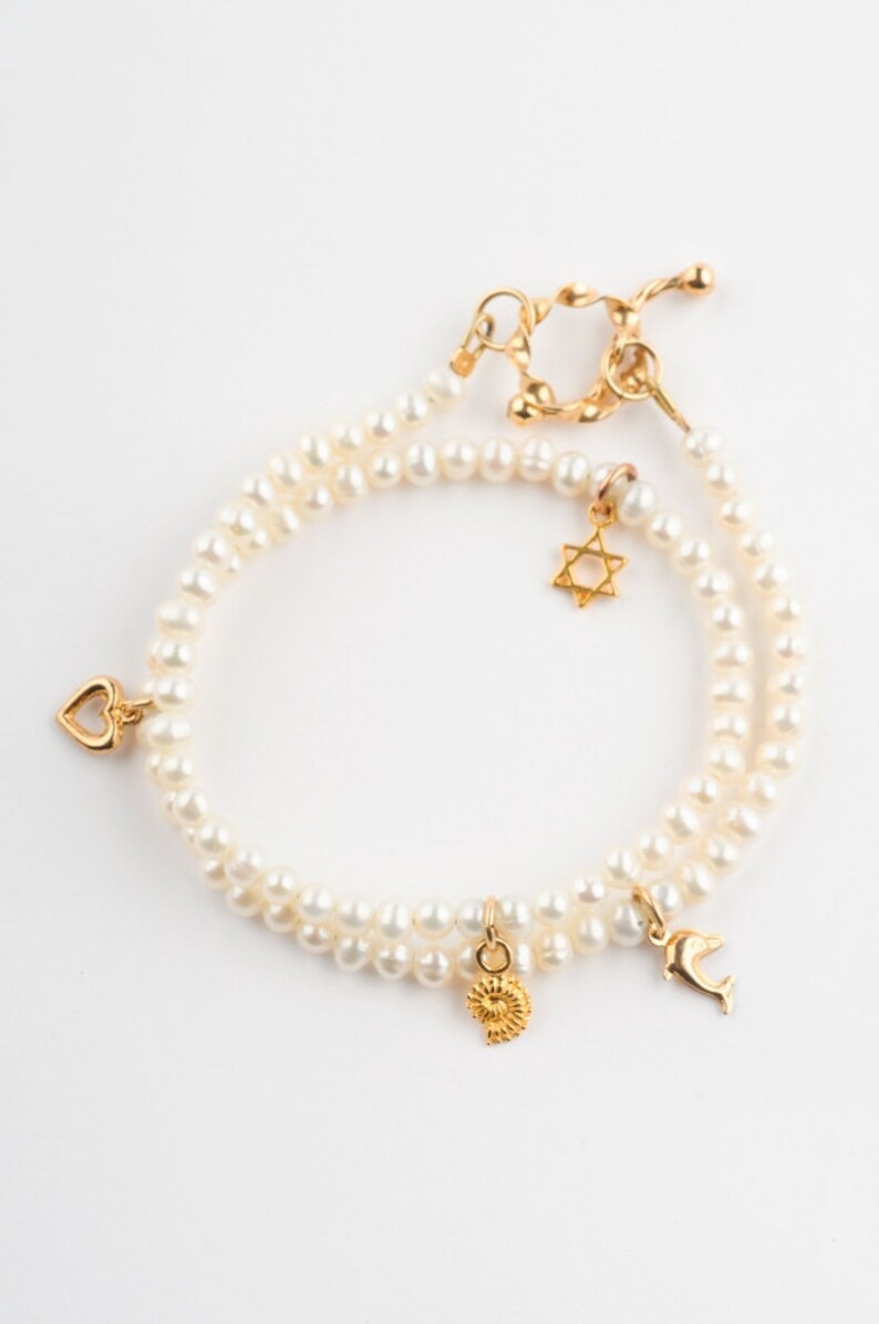 May include: A double strand pearl bracelet with gold charms. The charms include a heart, a star of David, a seashell, and a dolphin.