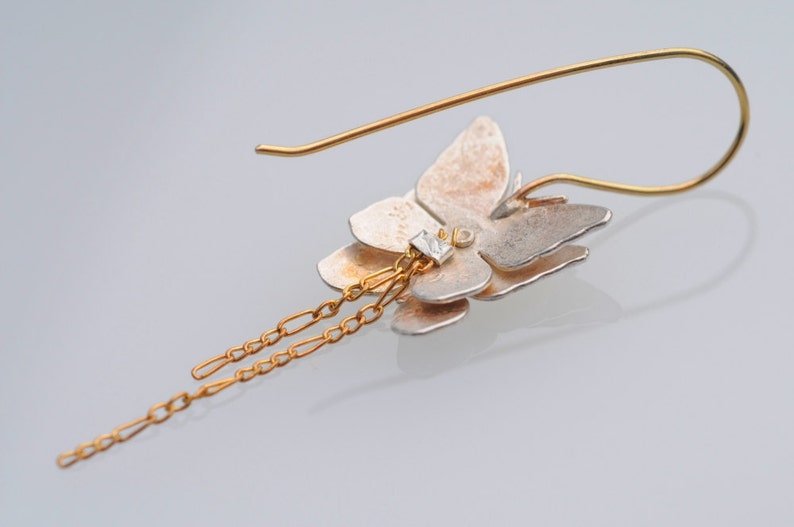 May include: A silver and gold butterfly earring with a dangling chain. The butterfly has a clear stone in the center.