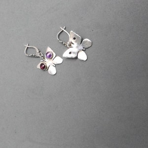 May include: A pair of silver butterfly earrings with purple and red gemstones. The earrings are on a gray background.
