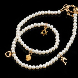 May include: Two pearl necklaces with gold charms. The larger necklace has a dolphin charm, a heart charm, and a gold clasp. The smaller necklace has a star of David charm and a spiral charm.