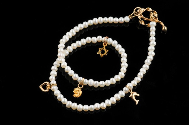 May include: Two pearl necklaces with gold charms. The larger necklace has a dolphin charm, a heart charm, and a gold clasp. The smaller necklace has a star of David charm and a spiral charm.