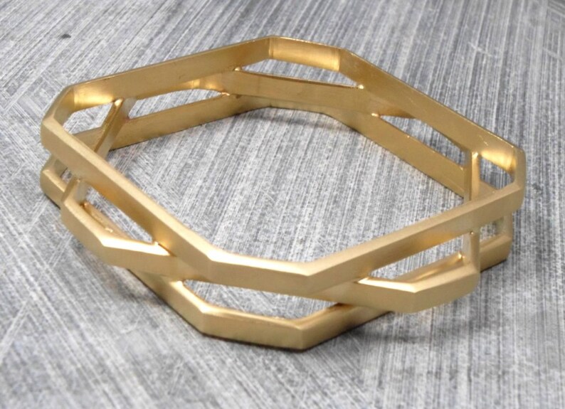 May include: Gold-toned metal bangle bracelet with a geometric design. The bracelet features an open, interlocking design with multiple hexagonal shapes. The bracelet is approximately 2.5 inches in diameter.