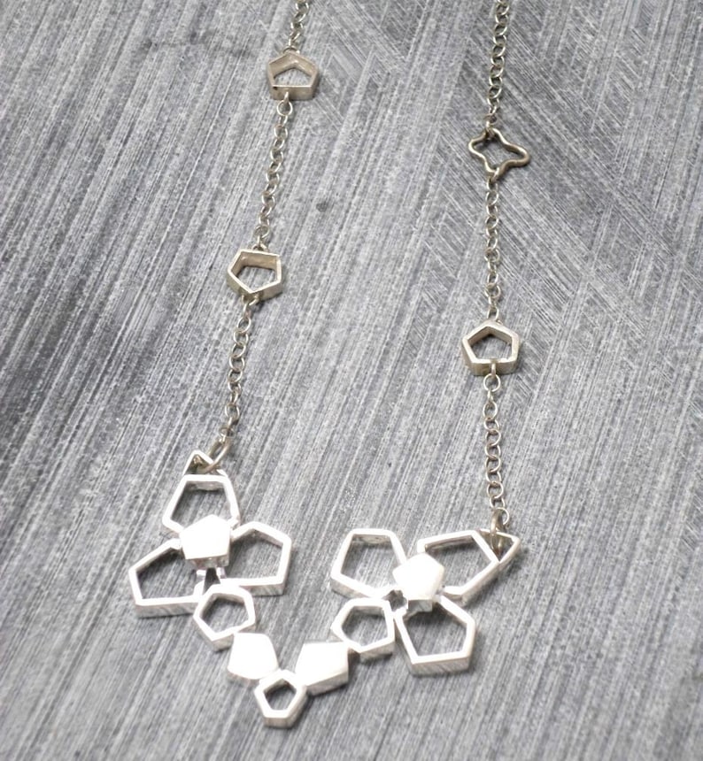 May include: A silver necklace with a geometric design. The necklace features a series of interconnected, open-frame shapes that resemble small, abstract flowers.