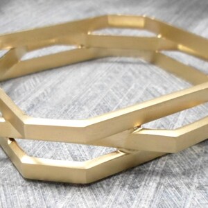 May include: Gold-colored geometric bangle bracelets. The bracelets have a unique, angular design with interlocking shapes. The metal has a matte finish, and the bracelets are displayed on a textured, gray surface. The overall aesthetic is modern and minimalist.